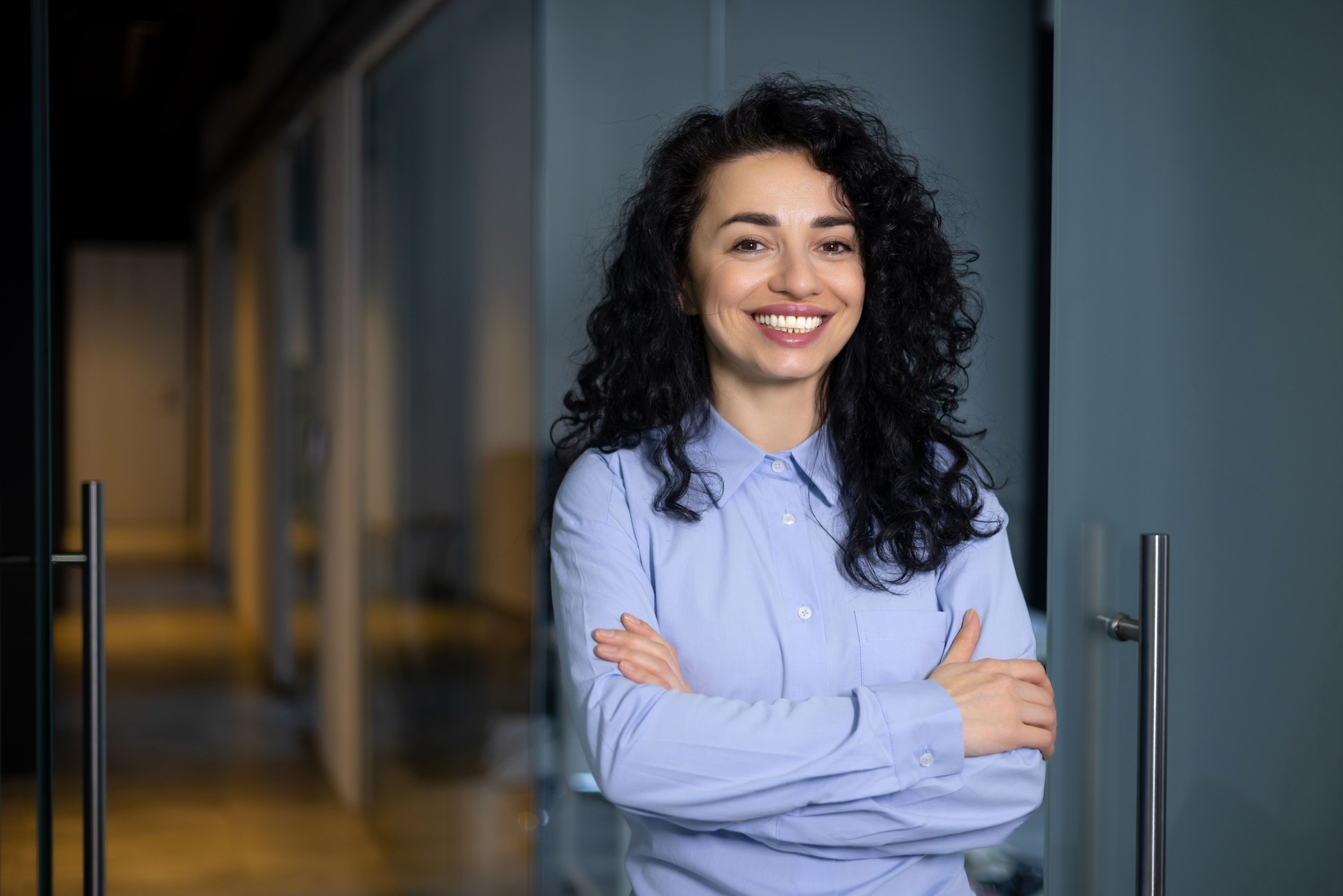 Portrait of happy and successful business woman, boss in blue shirt smiling and looking at camera inside office with crossed arms, Hispanic woman with curly hair in corridor